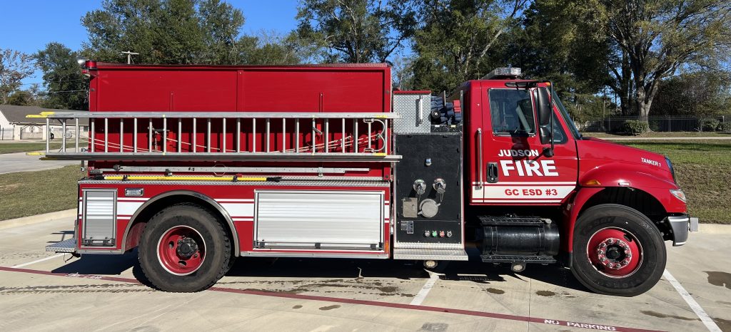 Side view of a red fire engine marked "Judson Fire" parked on a concrete surface with clear skies in the background. A ladder is mounted on the side, and trees are visible in the distance.