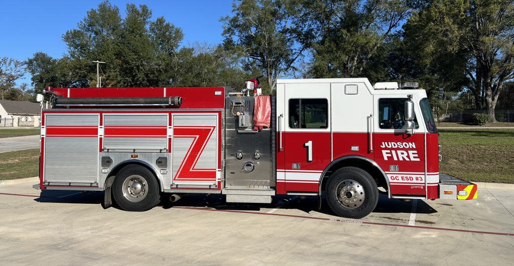 A red and white fire truck with "Judson Fire" written on the side is parked on a paved area. The truck has a large hose, compartments, and the number 1 displayed. Trees and a building are in the background under a clear blue sky.