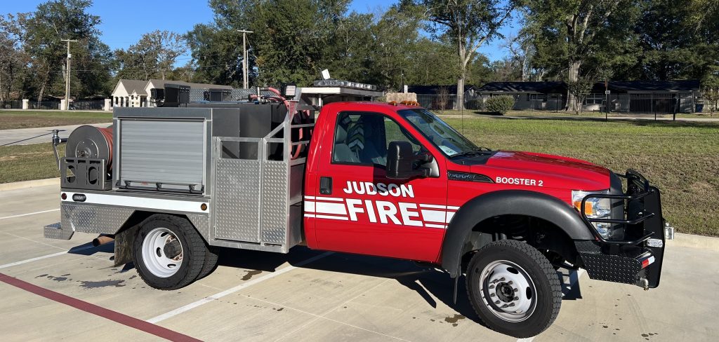 A red fire truck labeled "Judson Fire Booster 2" is parked on a paved area. It has a silver equipment compartment and hose reel. Trees and buildings are visible in the background under a clear blue sky.
