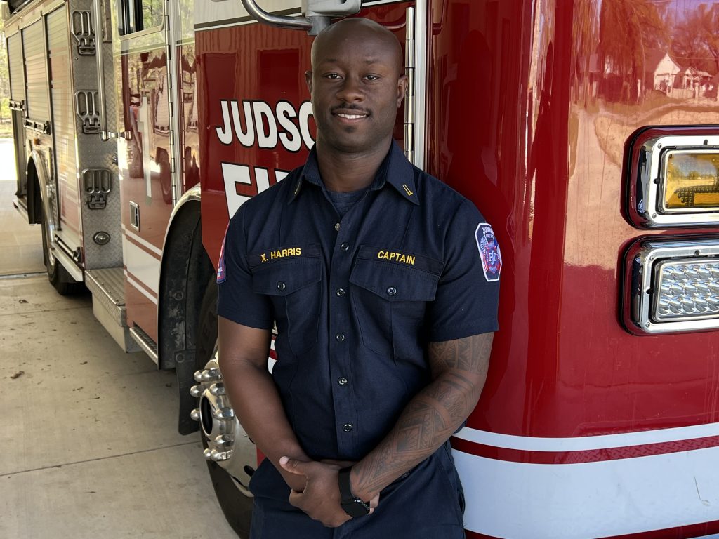 A firefighter in uniform stands confidently in front of a red fire truck, with "Judson Fire" written on it. He wears a short-sleeve, buttoned navy shirt with "Captain" insignia and a sleeve patch. His arms are relaxed by his sides.