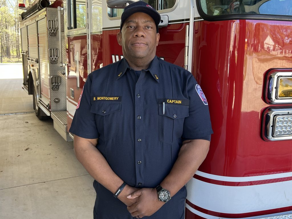 A firefighter in uniform stands in front of a red fire truck. He wears a navy blue shirt with patches and a black cap. The fire truck is parked in a station, and sunlight is visible in the background.