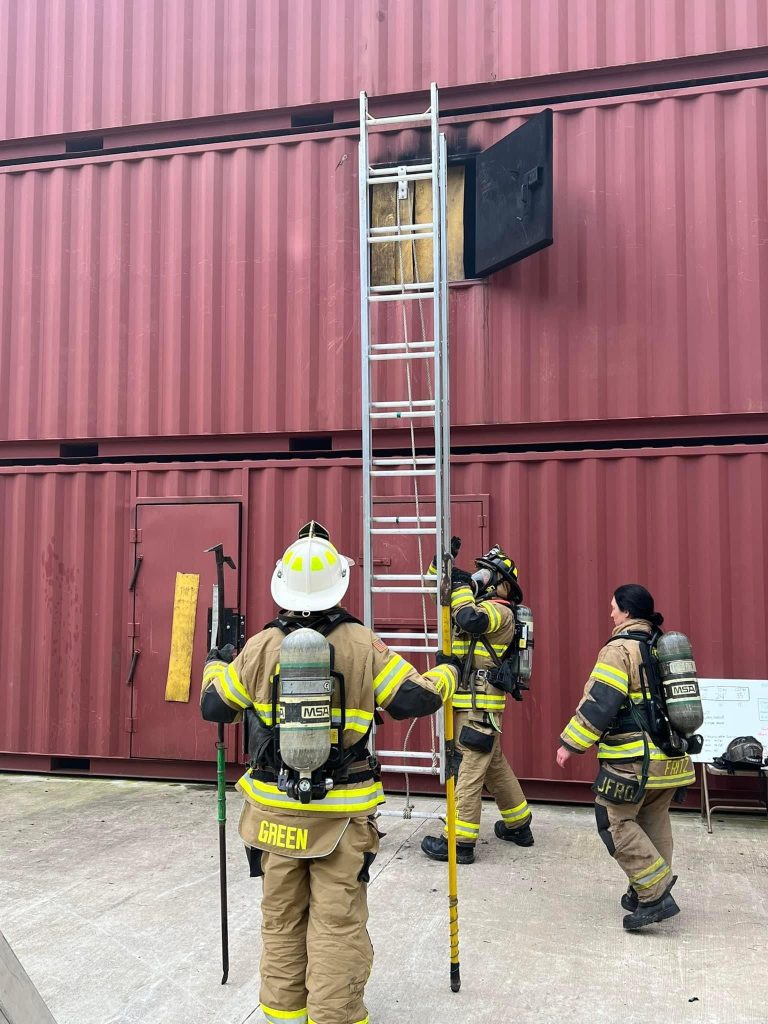 Three firefighters in gear stand by a ladder leaning against stacked red shipping containers. One is climbing, while the others hold tools and look up. A black door on the container is open near the top of the ladder.