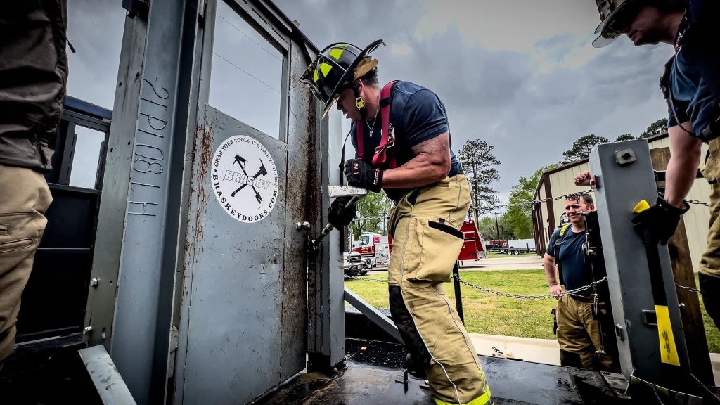 A firefighter wearing a helmet and gear uses a tool to force open a door during a training exercise. Another firefighter observes nearby. A logo on the door reads "Brothers in Battle." Overcast sky and trees form the background.
