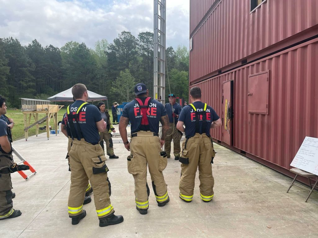 A group of firefighters in uniform and fire-resistant pants stand near a training building. They are engaged in discussion, with some facing the building. A forested area and a few scattered equipment pieces are visible in the background.