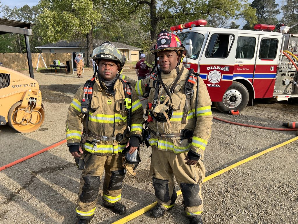 Two firefighters in full gear stand near a fire truck outside. They wear helmets, reflective suits, and equipment. A construction vehicle and trees are visible in the background under a clear sky.