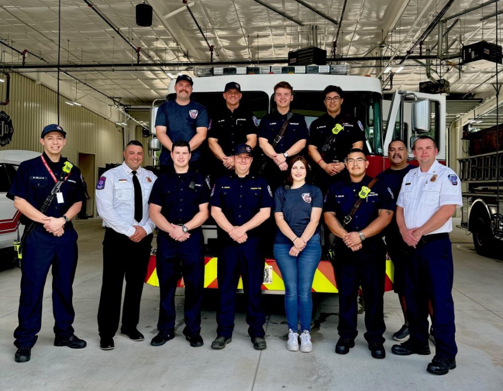 A group of eleven firefighters, both men and women, stand and sit in uniform in front of a fire truck inside a fire station. The setting is bright and the atmosphere is friendly.