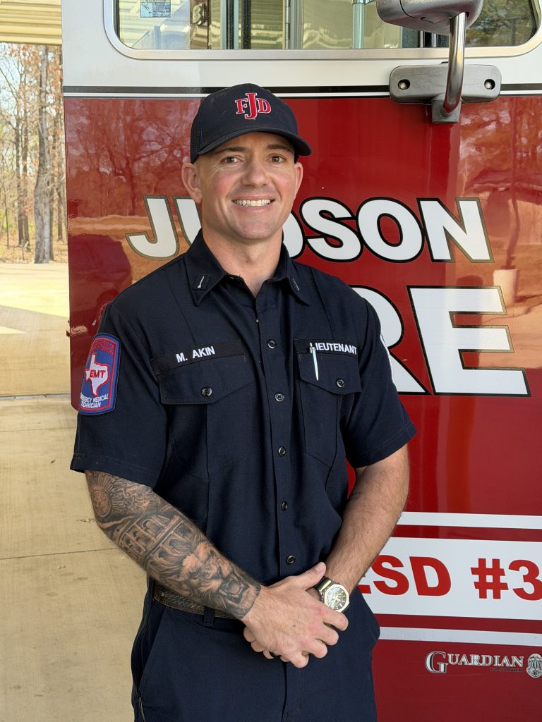 A firefighter in uniform stands smiling in front of a red fire truck. He wears a black cap with "FJD" on it, and his uniform displays the name "M. Akin" and a patch reading "EMT." Trees are visible in the background.