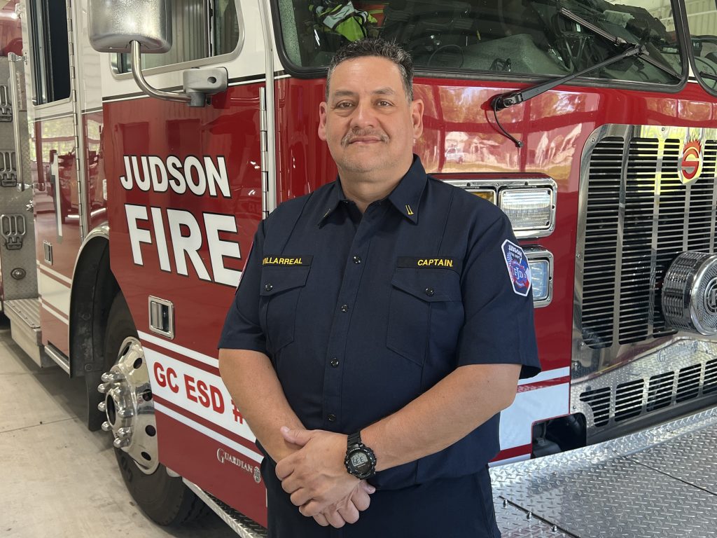 A firefighter in uniform stands in front of a red fire truck with "Judson Fire" written on it. The firefighter has a badge labeled "Captain" and is smiling at the camera.